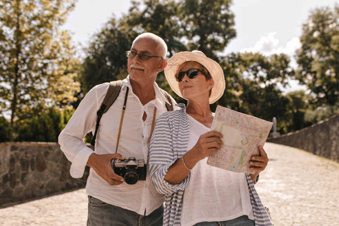 Short haired lady in hat, cool sunglasses and striped blue clothes holding map and posing with man in glasses and white shirt with camera in park. propósitos de viaje